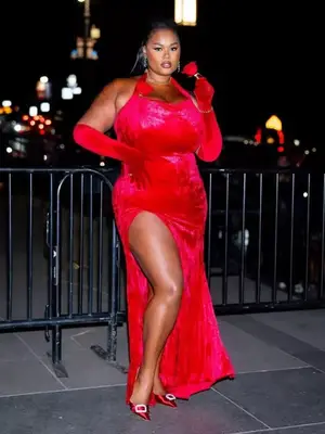Model Precious Lee is standing outside wearing a red velvet halter dress with matching red gloves holding a red rose. 
