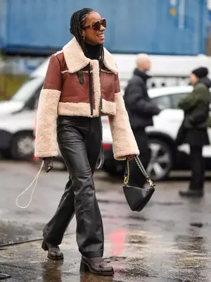 Woman wearing a brown leather coat with shearling trim, black leather pants, a triangle black bag, boots, and sunglasses