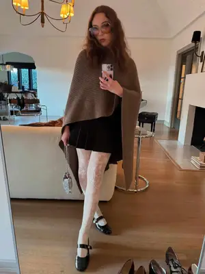 Laufey posing for a photo in a brown cape, black skirt, white lace leggings, and black patent flats