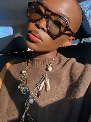 Woman wears tinted aviator sunglasses, feather charm necklace, and brown sweater