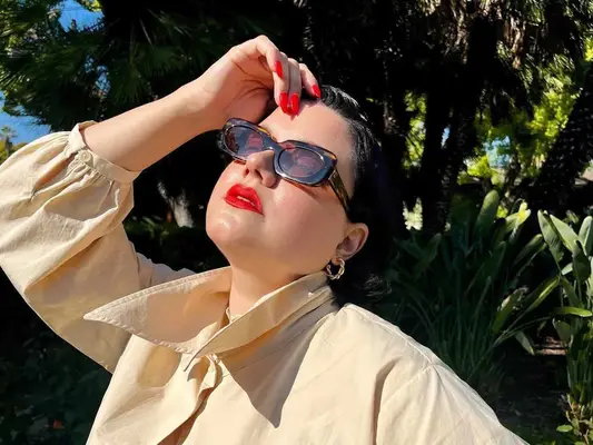Woman wearing tortoise rectangular sunglasses, button-down shirt, hoop earrings, and red lips and nails