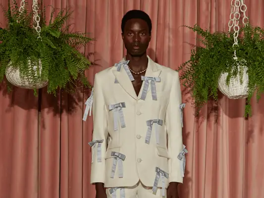 Model wearing white suit with powder blue bow appliques while standing near hanging plant baskets