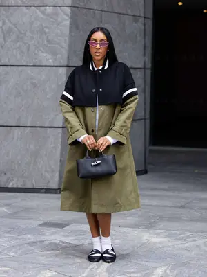 Tayshia Adams poses for a photo while wearing a green and navy trench coat, black ballet flats, socks, and a Longchamp Le Roseau bag