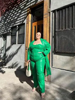 Abby Bible wears a green leather outfit comprising a corseted tube top, pants, and a trench coat plus white handbag, heeled sandals, and sunglasses