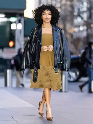 Yara Shahidi wears an oversized black moto jacket, gold dress with ruffled tiered skirt, brown pumps, and gold jewelry