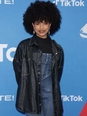 Kathryn Rodriguez wears a silky black button-down, black mock neck top, and denim overalls