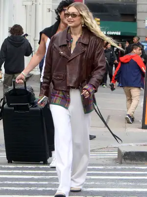 Gigi Hadid wears a brown leather jacket, plaid shirt, white trousers, boots, and sunglasses