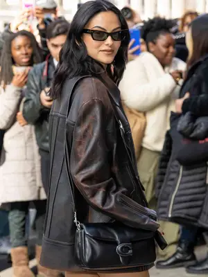 Camila Mendes wears a brown leather jacket, black Coach shoulder bag, angled sunglasses, and wavy hairstyle