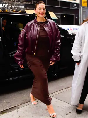 Ashley Graham wears a wine-colored leather bomber jacket, brown maxi dress, open-toed heels, and gold hoop earrings