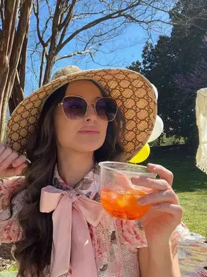 Kacey Musgraves wears a sunhat, pink floral blouse with ribbon, sunglasses, and holds an Aperol spritz