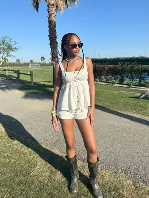 Woman wearing white babydoll tank top, lightweight knit shorts, biker boots, black sunglasses, and layered necklaces and bracelets