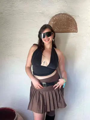 Barbie Ferreira wears a black crop top with plunge neckline, brown pleated skirt, black belt, and shield sunglasses