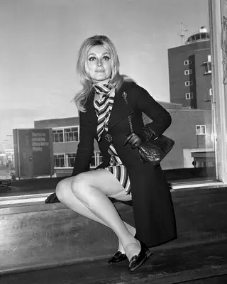 Sharon Tate at the airport wearing a striped dress with a long coat, black handbag, gloves, and buckled shoes