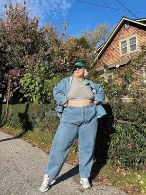 Abby Bible wears a light blue denim jacket, gray cropped t-shirt, wide leg jeans, sneakers, green baseball cap, and sunglasses