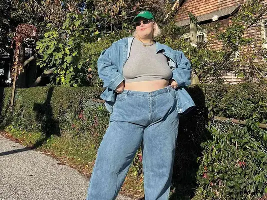 Abby Bible wears a gray cropped t-shirt, denim jacket, jeans, green baseball cap, and sunglasses