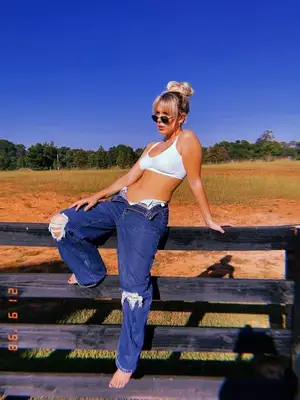 Millie Bobby Brown wears unbuttoned jeans, a white bra top, and sunglasses while sitting on fence