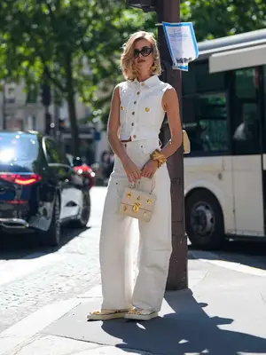 Xenia Adonts wears black sunglasses, gold Schiaparelli earrings, white and gold button up tailored vest, gold bracelet, white loose denim jean pants, beige sneaker shoes, white with gold detail Schiaparelli leather bag
