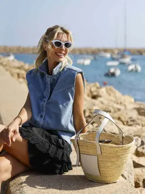 Annika Gassner, wearing a black skirt, a sleeveless denim jacket, white sunglasses, and a raffia handbag