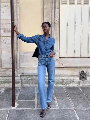 Sylvie Mus wearing a denim button-down top, light straight leg jeans, a black handbag, and black open-toed shoes. 
