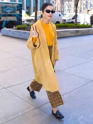 Zoe Kravitz wears a yellow crewneck top, suede coat, plaid pants, sunglasses, and loafers