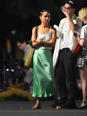 Zoe Kravitz wears a white tank top, green slip skirt, neutral sandals, and a shoulder bag