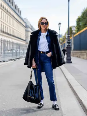 Woman wearing denim jacket, jeans, white t-shirt, leather shearling coat, oversized Chanel bag, loafers, socks, and sunglasses