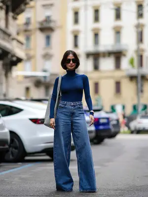 Woman wearing blue turtleneck top, extra wide leg jeans, a woven shoulder bag, and sunglasses