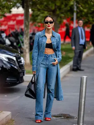 Woman wearing long denim trench coat, straight leg jeans, scarlet heels, oversized black handbag, and sunglasses