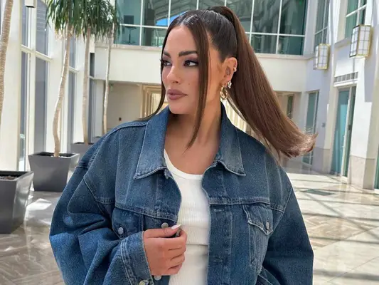 Ashley Graham wears a white ribbed tank top, denim jacket, and high ponytail hairstyle with face-framing pieces