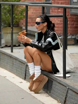 Bella Hadid wears mini Ugg boots, white crew socks, an oversized leather jacket, graphic tee, and shield sunglasses