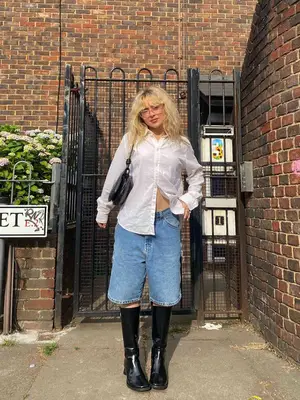 Sabrina Carpenter wears a white button-down shirt, denim Bermuda shorts, square-toe knee-high chunky boots, a black shoulder bag, and glasses