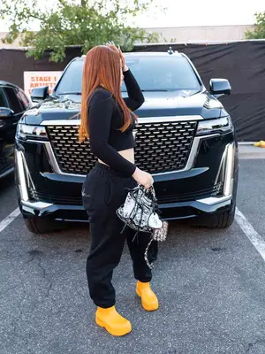 Ice Spice wearing black cropped long-sleeve shirt, black sweatpants, patterned handbag, and yellow rubber chunky boots