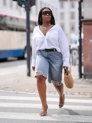 Person wearing a button-down white shirt, denim shorts, sunglasses, layered necklaces, and heels
