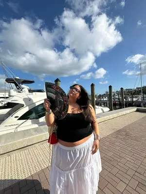 Model Priscilla Del Castillo is standing outside next to a boat holding a black fan. They are wearing a black tube top, a white maxi skirt, and black sunglasses. 