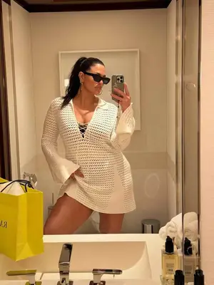 Model Ashley Graham is standing in front of the mirror wearing a white crochet long sleeve dress, black sunglasses, and silver hoops. 