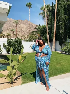 Influencer, @itsmekellieb is standing outside in front of palm trees wearing a blue paisley print long-sleeve tie front top with matching maxi slit skirt. 