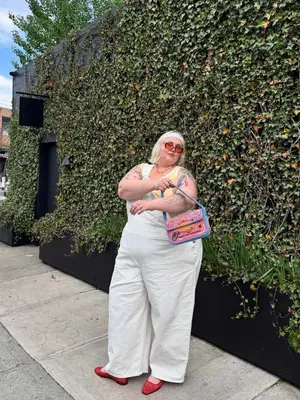 Influencer @theabbybible is standing outside a wall of moss wearing a yellow and white stripe tank top, white overalls, and a pink and blue fish bag with red ballet flats
