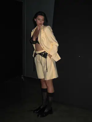 Model @lcchan is standing in the dark wearing a butter yellow suit jacket and shorts with a black bra under and a black lace gathered over. 