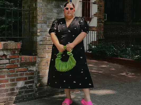 Jessica Torres wears a black and white sundress, green ruched handbag, pink slide sandals, and peach sunglasses