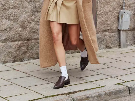 Close-up of woman wearing dark brown loafers, white crew socks, tan pleated skirt, and longline beige coat