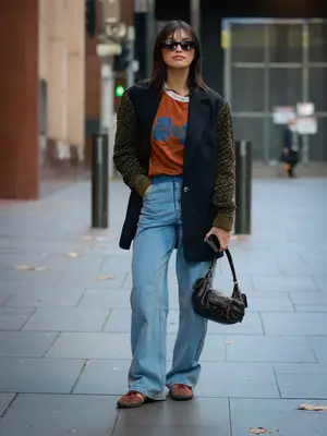 Sarah Ellen wears a collared jacket with knit sleeves, orange collared t-shirt, jeans, and pocketed black bag