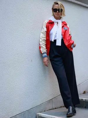 Woman with varsity jacket, white shirt, sweater tied around neck, black wide leg pants, boots, and sunglasses