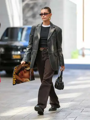 Hailey Bieber wears a black shirt with white collar, brown tailored pants, and a structured jacket