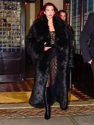 Dua Lipa wearing a large black fur jacket, sheer lace dress, and black boots