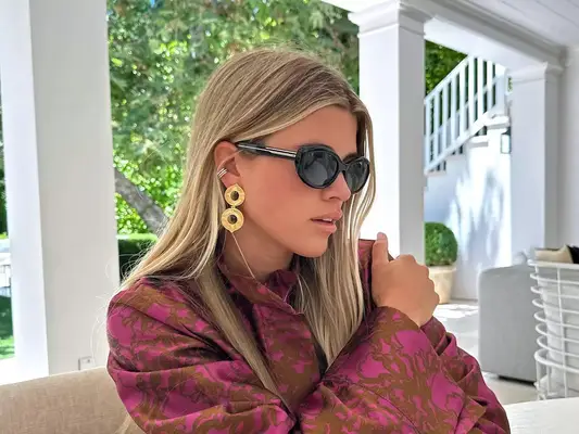 Sofia Richie Grainge wears a pink and brown patterned top, gold earrings, and black quiet luxury sunglasses