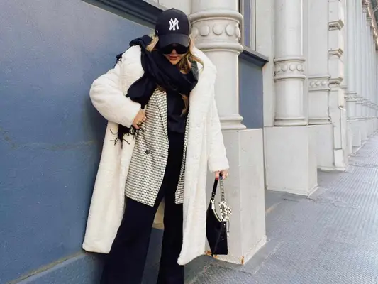 Audree Kate Lopez wears a white fluffy coat, checked blazer, scarf, New York Yankees hat, trousers, and black handbag