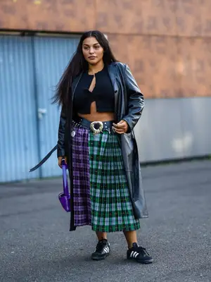 Paloma Elsesser wears a black cardigan, leather trench coat, purple and green tartan skirt, belt, black Adidas Sambas, and purple handbag