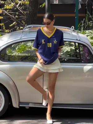 Woman wearing blue and yellow v neck jersey, ruffled cream mini skirt, white ballet flats, gold necklace, and brown sunglasses