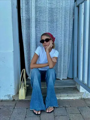 Woman wearing blue jeans, white tee, black sunglasses, yellow bag, red bandana, and flip flops