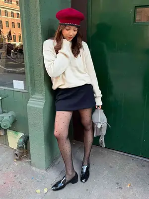 Laufey posing for a photo while wearing swiss dot tights, a black mini skirt, white sweater and red beret